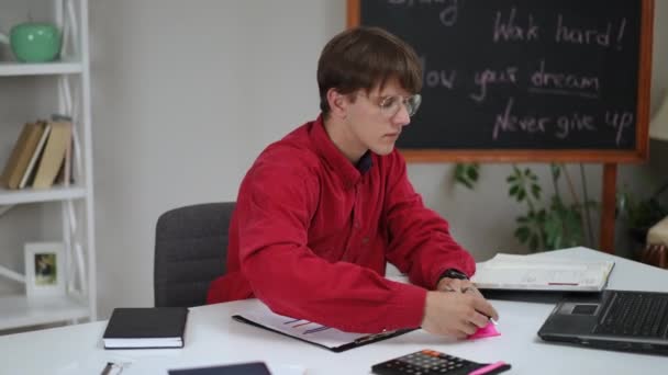 一个戴眼镜的年轻人用钢笔在粉红的便条上写字 并贴在桌子上 一个男人拿着一个带有图表的剪贴板 微笑着在笔记本电脑的摄像机前展示 它就站在屏幕上 — 图库视频影像