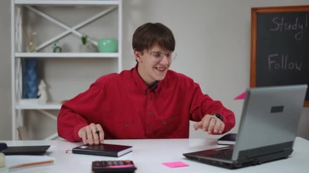 一个戴眼镜的年轻人在家里坐在桌子旁 一边用笔记本电脑一边聊天 一边笑着 那家伙把一个开着的笔记本电脑拉向他 然后摘下眼镜 — 图库视频影像