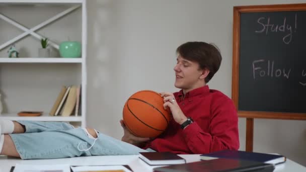 男はテーブルの上の足で椅子に座り 家の屋内で座っている間バスケットボールで遊ぶ笑顔 — ストック動画