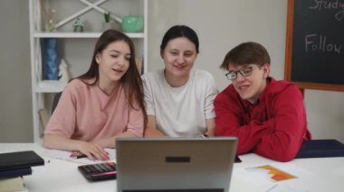 Öğrenciler bir dizüstü bilgisayar ve internet kullanarak iletişim kurarlar. Bir adam ve iki kız dizüstü bilgisayarın kamerasına bakar, gülümser, ellerini sallar ve bir not defterinden notlar okur, oturma odasındaki bir masada otururlar.