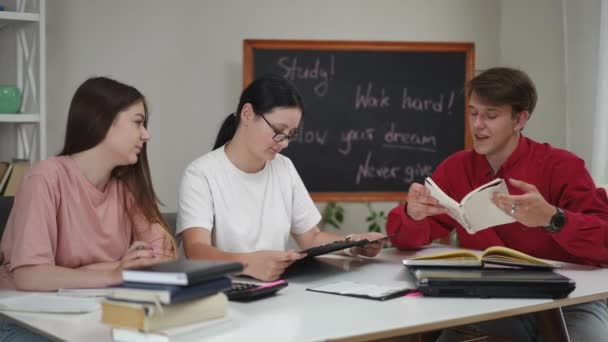 一个穿红衫的男人坐在一间房子的客厅里的桌子旁边 给两个女孩读了一本书 这家伙笑着把他的手指放在书页上 学生们一起学习准备考试 — 图库视频影像