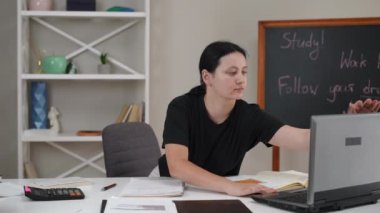 Bir kız öğrenci masada oturuyor, bilgisayarını elleriyle açıyor, ders notlarına bakıyor ve not defterine kalemle yazıyor. Enstitüde sınavlara hazırlanılıyor