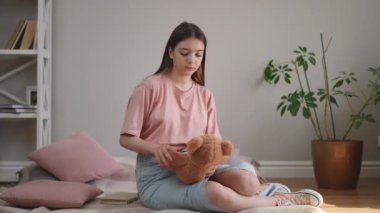Uzun saçlı bir kız oyuncak ayıya sarılıyor ve düşünceli bir şekilde uzaktan bakıyor. Oturma odasının ahşap zemininde bir ekosenin üzerinde oturuyor.