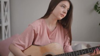 Yakın plan. Uzun saçlı bir kız elinde akustik bir gitar tutuyor ve gülümsüyor, parmaklarıyla telleri koparıyor, oturma odasındaki ahşap zeminde bir battaniyeye oturuyor.