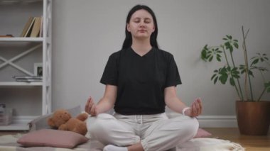Siyah tişörtlü genç bir kadın gözleri kapalı meditasyon yapıyor. Oturma odasındaki ahşap bir zeminde lotus pozisyonunda oturuyor. Rahatlamak için nefes alma egzersizleri. Yoga alıştırması.