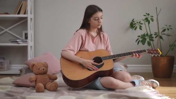一个长着长发的可爱女孩 手里拿着一把音响吉他 用手指心事重重地抓起琴弦 坐在客厅地板上的格子花上 — 图库视频影像