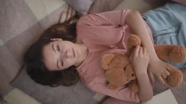 顶部视图 在客厅里 一个长头发的可爱而悲伤的女孩抱着一只泰迪熊躺在地上 头枕在枕头上 — 图库视频影像