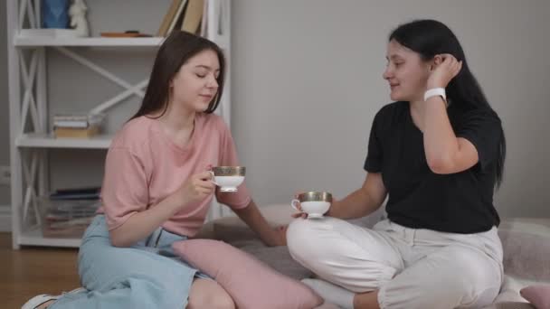 リビングルームの床で飲んでいるお茶 二人の女の子が話し 笑って モダンな家の部屋の毛布の上に床に座っている間 磁器ティーカップを手に握ります — ストック動画