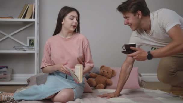 一个女孩坐在地板上的格子花上看书 那家伙带来了一个咖啡杯和茶托 并把它给了女孩 那人和那女孩坐在毛毯上 他们开始交谈 — 图库视频影像