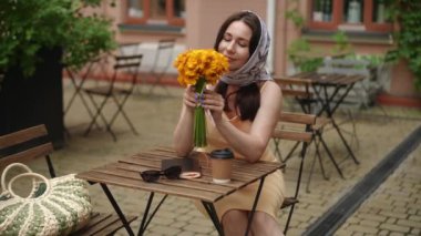 Ağır çekim. Başörtülü şık bir kadın, bir yaz günü şehir kafesinde bir masada otururken elinde sarı bir çiçek buketi tutuyor. Kadın bukete bakıyor, kokluyor ve...