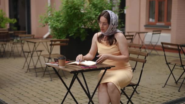 慢动作夏天的一天 一个戴眼镜 头戴头巾的时髦女人坐在城市室外咖啡馆的桌子边 一边用钢笔在笔记本上写字 那个女人关上笔记本 把她带走了 — 图库视频影像