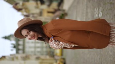 Dikey video. Ağır çekim. Şapkalı ve omuzlarında kadın ceketi olan sevimli bir kadın gülümseyen kameraya bakıyor. Bir kadın kameraya arkasını döner ve şehir caddesinde yürür.