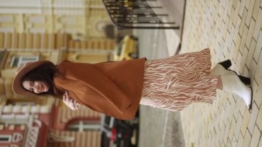 Dikey video. Ağır çekim. Dolgun şapkalı, omuzlarında kadın ceketi olan mutlu, şık bir kadın yaz günü bir caddede dikilirken dönüp duruyor. Gülümseyen güzel kadın