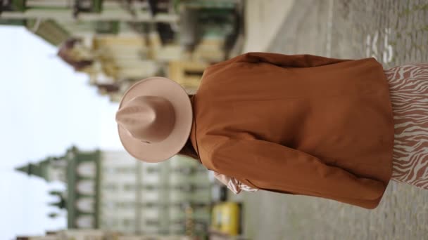 バーティカルビデオ スローモーション 彼女の肩に輝いた帽子と女性のジャケットの女性は カメラの背中に彼女の後ろに立っています 女性が振り返って 街の通りで立っているカメラを見ます — ストック動画