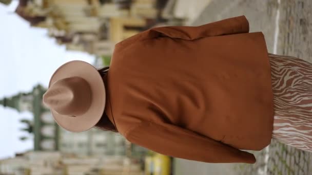 垂直录像 慢动作后视镜夏天的一天 一个戴着帽檐帽 肩上披着女式夹克的女人沿着城市街道走着 一个漂亮的女人转过头看着摄像机 — 图库视频影像