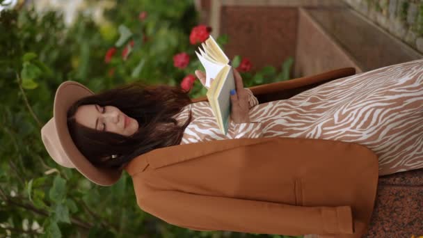 バーティカルビデオ スローモーション 彼女の肩に輝く帽子と女性のジャケットのスタイリッシュな女性は 熱狂的に本を読んでおり 彼女は夏に市立公園に立っている — ストック動画
