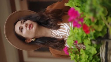 Dikey video. Ağır çekim. Yakın plan. Dolgun şapkalı ve kadın ceketli şık bir kadın dışarıda çiçek dolu arka planda dikilirken gülümsüyor ve bakıyor.