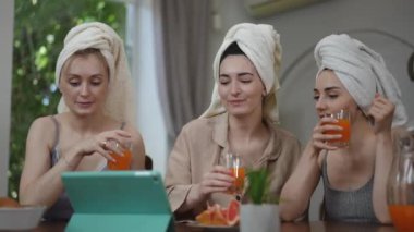 Kafasında havlu olan üç güzel kız gözlüklerinden portakal suyu içerler ve masanın üzerinde duran tabletin ekranına bakarlar. Kızlar kahvaltı yapar, gülümser ve güler, masada otururlar.