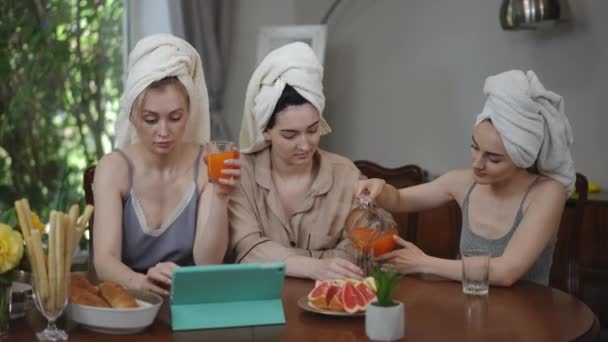彼らの頭にタオルが付いている3人の少女は 朝のリビングルームのテーブルに座っている間 ガラスジャグからオレンジジュースを注ぎます — ストック動画