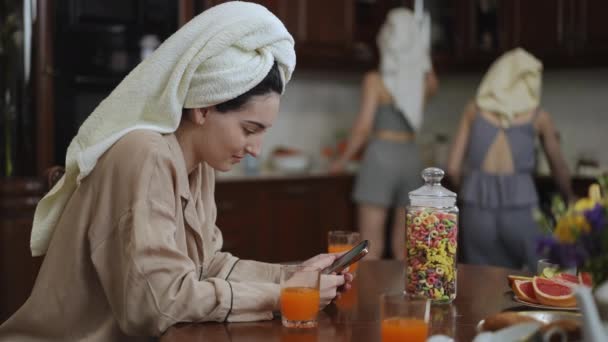 漂亮的女人头上拿着毛巾在一间现代化的房子的厨房里准备早餐 一个女孩坐在桌旁 看着手机屏幕 — 图库视频影像