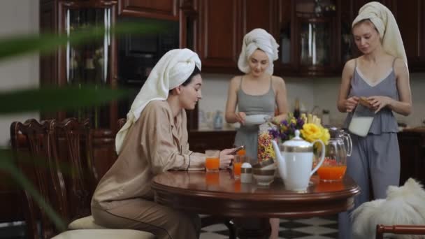 早上准备早餐 漂亮的女人头上拿着毛巾在一间现代化的房子的厨房里准备早餐 一个女孩坐在桌旁 给她的朋友们看她的屏幕 — 图库视频影像