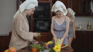Kızlar kahvaltıda taze sebzelerden salata hazırlar, modern bir mutfakta dururlar. Kafalarında havlu olan genç kadınlar roka 'yı cam bir kaseye koyup taze sebzeleri mutfak ile kesiyorlar.