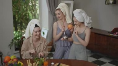 Kafalarında havluyla dans eden üç güzel kız arkadaş ellerinde mutfak gereçleri, modern bir mutfakta kahvaltı hazırlarken neşeyle dans ediyorlar. Kadınlar eğleniyor, gülüyor ve gülümsüyor.