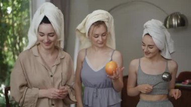 Kafalarında havlu ve ellerinde mutfak gereçleri olan üç genç bayanın neşeli dansı. Kadınlar eğleniyor, gülümsüyor ve gülüyor.