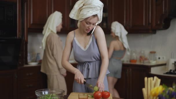 在前景中 一位头上拿着毛巾的年轻女子站在厨房里 一边用手机交谈 一边用菜刀在木板上切新鲜蔬菜 — 图库视频影像