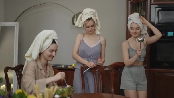 两个头上拿着毛巾的女孩手拿着厨房用具在一个现代化的厨房里跳舞 第三个女孩坐在桌旁搅拌蔬菜沙拉 快乐的女人在准备 — 图库视频影像