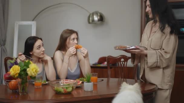 2人の女の子がリビングルームのテーブルに座り 新鮮な野菜サラダとフルーツを朝食に食べています 3人目の女の子が熱いピザでテーブルに近づき 友達にそれを提供する — ストック動画