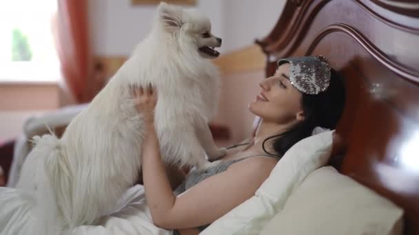 一位漂亮的年轻女子躺在床上 手里拿着一只白色的波美拉尼亚斯皮茨 女孩笑着看了看狗 — 图库视频影像