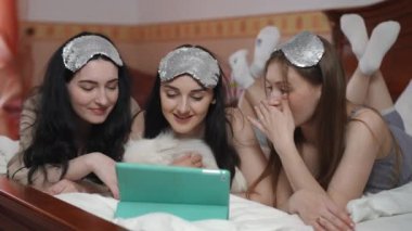 Üç gülümseyen genç kadın, kafalarında uyku maskeleri, ve beyaz bir Spitz, yatağa uzan ve tablet ekranına bak.