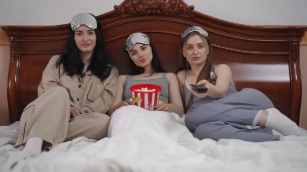 三位漂亮的年轻女性头戴睡眠面具 坐在卧室的床上 手里拿着一大杯塑料爆米花 看着摄像机 那女孩有一台电视遥控器 — 图库视频影像