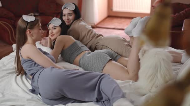 这个女孩和她的女朋友躺在卧室的床上 讲述了一个迷人的故事 年轻的女人头上戴着银色的睡眠面具 一只白色的斯皮茨犬在认真地听着 — 图库视频影像