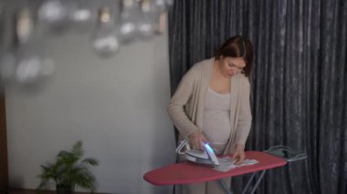 Evde kıyafetleri olan hamile bir kadın evinin oturma odasında dururken ütü masasında ütü ütülüyor. Kadın elindeki bebek tişörtüne bakar ve başını sallar.