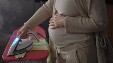 Yakın plan. Tanımlanamayan hamile bir kadın ütü masasındaki bebek kıyafetlerini ütülemek için buharlı ütü kullanır ve diğerini içeride dururken karnının üstünde tutar.