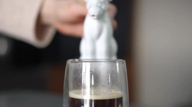 Yakın plan. Bir kadın, cam bir kahve bardağına inek şeklinde bir porselen kabın taze kahvesiyle süt döker.