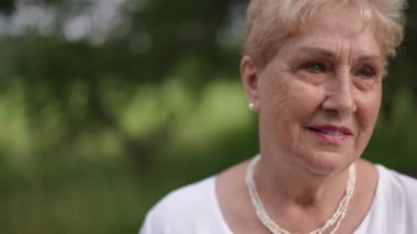 Yakın plan. Düşünceli yaşlı kadın bir yaz günü parkta dikiliyor. Yaşlı bir kadın gülümseyerek kameraya bakar.