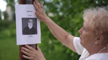 Yakın plan. Yaşlı bir kadın, şehir parkındaki bir direkte kaybolan yaşlı bir adam hakkında gazete ilanı yayınlıyor. Kadın tebligattaki fotoğrafa bakarken üzgün ve konuşuyor.