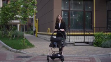 Bir kadın, çok katlı bir kompleksin avlusunda yaya geçidinden bebek arabasını iterken görülebilir. Bebek arabası sorunsuzca hareket ederken, elinde cep telefonu olan kadın etrafa bakıyor.