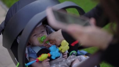 Yakın plan. Çocuk bebek arabasında yatıyor ve ağlıyor. Tanınmayan bir kadın, bir yaz günü elinde cep telefonuyla dışarıda beklerken bebeğe emzik veriyor..