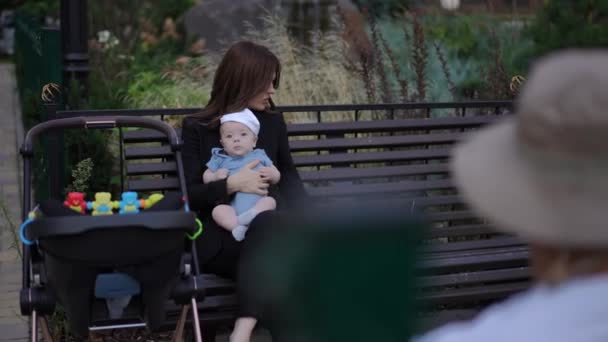 ショットオーバーショット 女性は彼女の腕に赤ん坊を抱え 屋外の夏の日にベンチに座っている間ノートパソコンで働きます — ストック動画