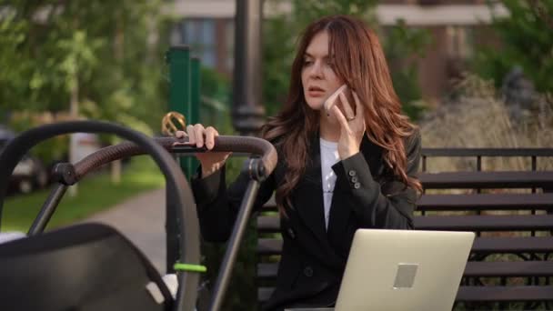 时髦的女商人坐在长椅上 带着敞着的笔记本电脑 摇晃着婴儿车 用手机交谈 妇女在产假期间外出工作 — 图库视频影像