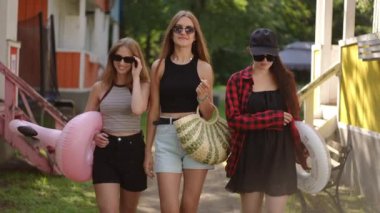Ağır çekim. Güneşli bir yaz gününde güneş gözlüklü üç kız dinlenme merkezinden gülümsüyor. Kız arkadaşlar kumsala güneşlenmeye ve havuzda yüzmeye giderler.