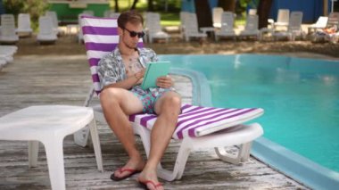 Ağır çekim. Güneş gözlüklü genç bir adam güneşli bir yaz gününde açık havuzun yanında plaj şezlongunda otururken elinde tabletle rahatlıyor. Adam bakar ve parmağını kaydırır.