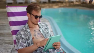 Ağır çekim. Güneş gözlüklü bir adam, güneşli bir yaz gününde yüzme havuzunun yanında plaj şezlongunda otururken elinde tabletle poz veriyor. Adam güneş gözlüğünü çıkardı ve baktı.