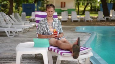 Ağır çekim. Genç bir adam tablet ekranına bakar ve kokteyl bardağından kokteyl içerken güneşli bir yaz gününde yüzme havuzunun yanında bir plaj şezlongunda oturur.