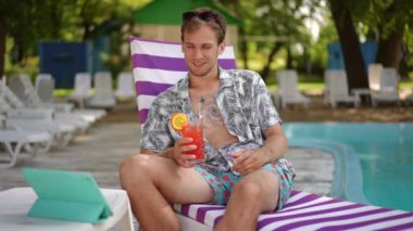 Ağır çekim. Bir adam internetten tablet kullanarak iletişim kuruyor ve açık havada güneşli bir yaz gününde plaj şezlongunda otururken bardaktan kokteyl içiyor.