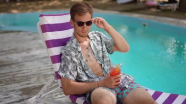 Ağır çekim. Bir adam kameraya poz veriyor ve kokteyl bardağından kokteyl içiyor, güneşli bir yaz gününde havuz kenarında bir plaj şezlongunda rahatça oturuyor. Bir adam gülümserken